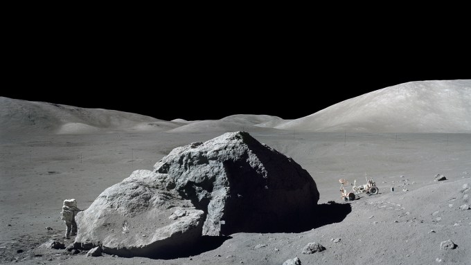 A black and white image of Apollo 17 astronaut Harrison Schmitt standing next to a lunar boulder on the moon.