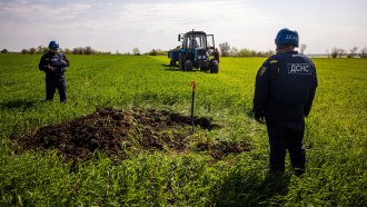 a team working to destroy an unexploded missile. Two people stand around a small post in a dug-up portion of field, with a blue vehicle in the distance