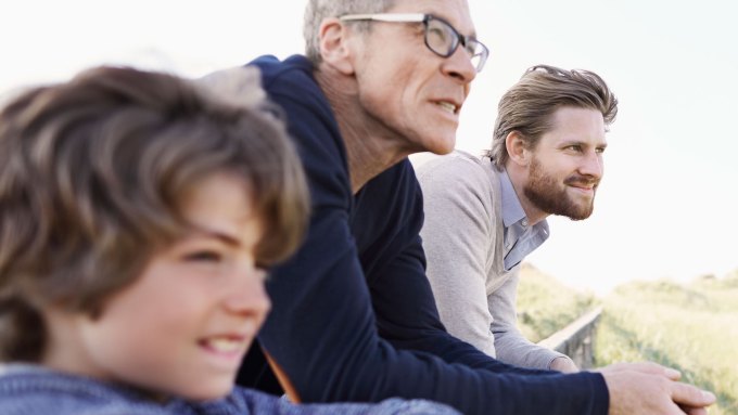 In this stock photo portraying a white multigenerational family, a young boy, older man and a middle-aged man lean on a fence near what looks like dunes and are all looking out toward the horizon.