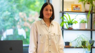 Andrea Gomez is standing in an office, softly smiling and staring at the camera. She has dark hair that comes just above her shoulders and is wearing off-white button-up jacket.