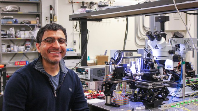 Bhavin Shastri stares into the camera while smiling widely. He's sitting in front of a laser table that has a microscope and other optical equipment on top. He is wearing a half-zip navy sweatshirt over a gray t-shirt. He is also wearing glasses.