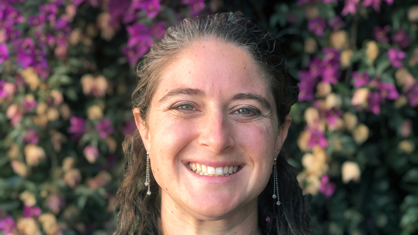 Jacqueline Gerson is staring at the camera with a wide smile. She has her dark brown hair pulled back in a half-up half-down. She's wearing earrings where each one has two spheres dangling on chains. The background is blurry, but it looks like she's standing in front of white and purple flowers.